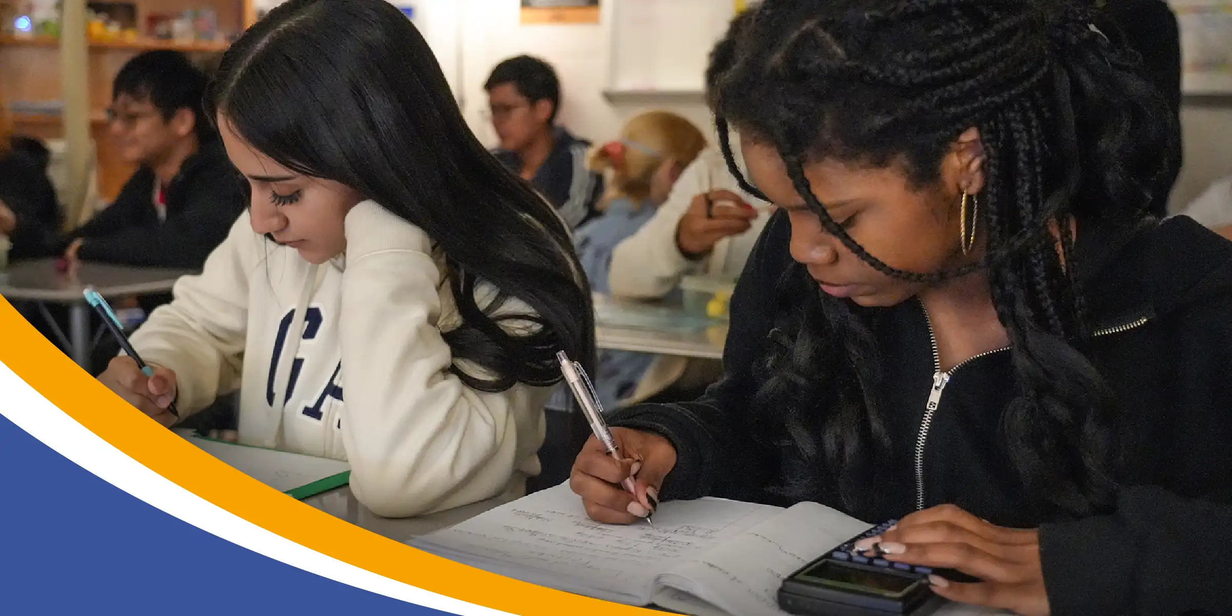 Two students in a classroom concentrate on their work, writing in notebooks at a desk.