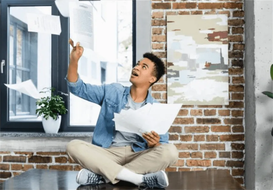 A young man sitting cross-legged on the floor catching sheets of paper that are falling from above