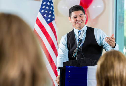 A person standing at a podium speaking before a crowd with a U.S. flag and balloons behind them