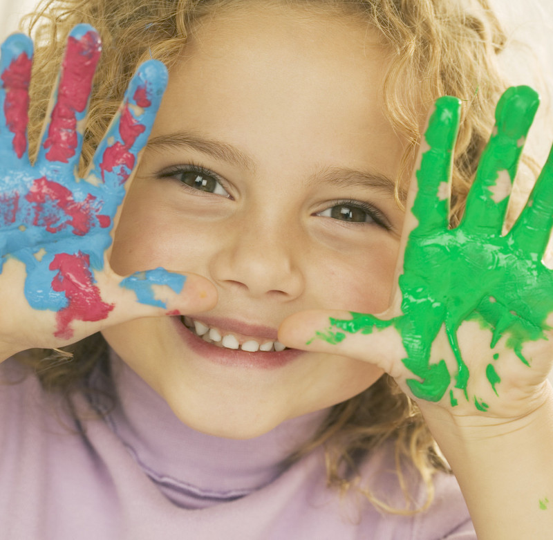 A toddler girl showing her hands covered in paint. One hand has green paint and the other hand had red and blue paint