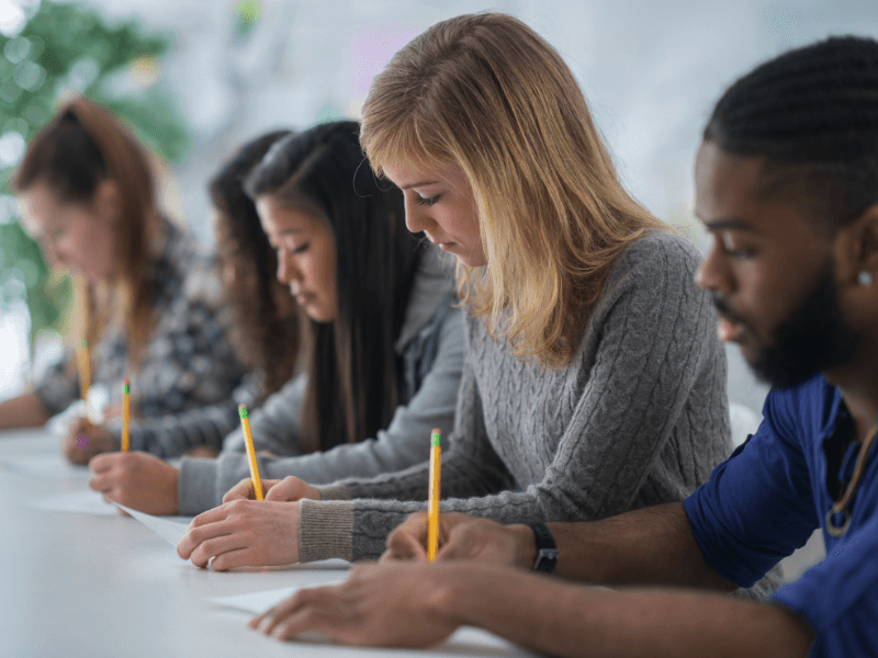 College Entrance Testing webpage banner with photo of a mouse on a standardized testing form Five students sitting at a desk taking a test with pencils