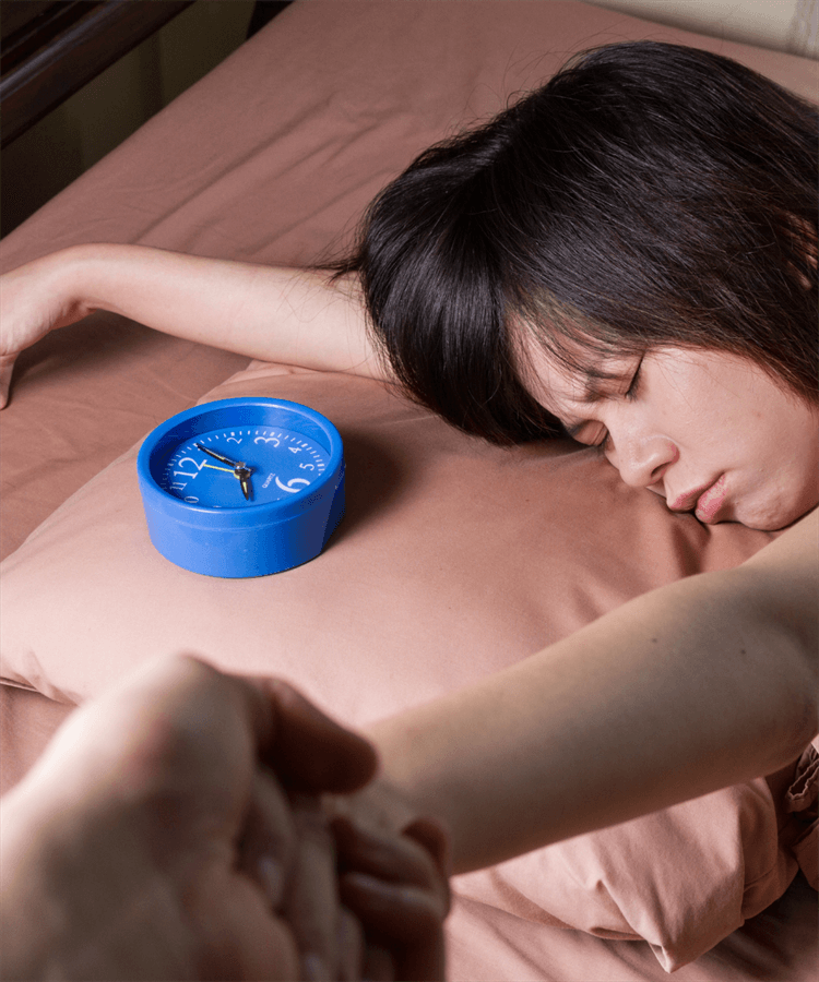 girl oversleeping in bed with alarm clock next to her head and someone reaching out to wake her up